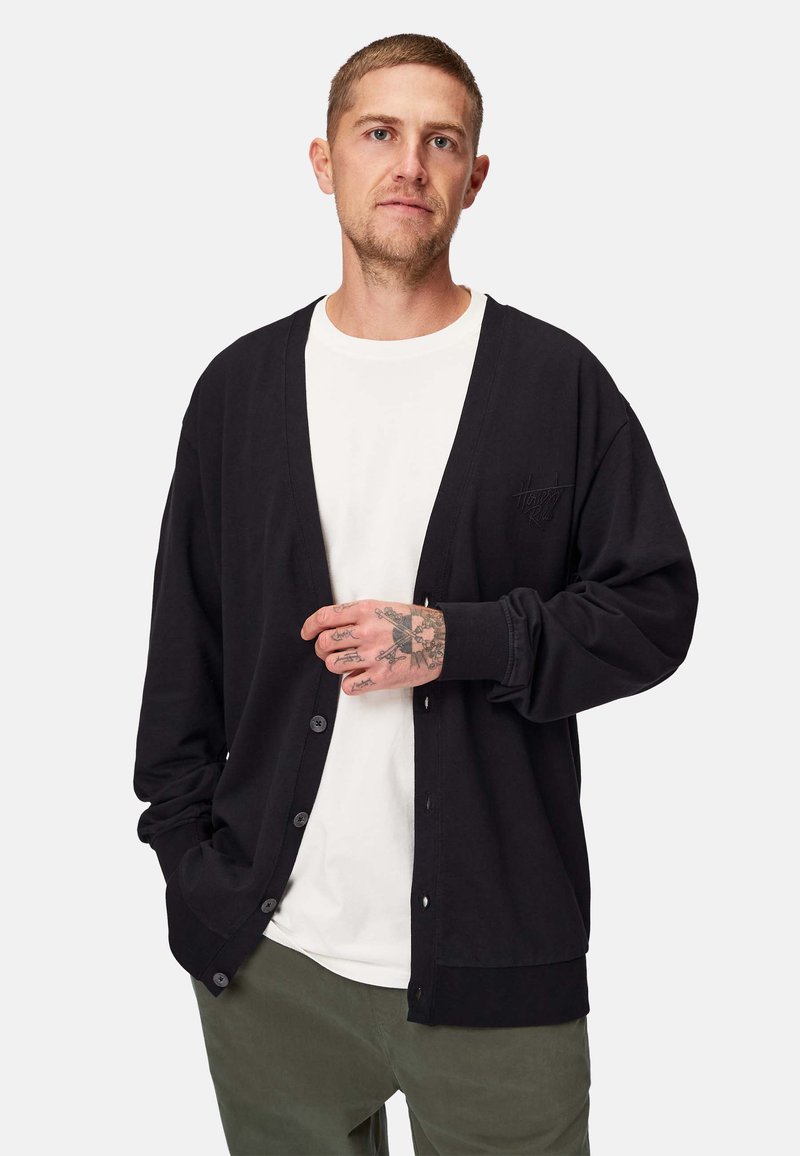 Sort cardigan lavet af blødt stof, med V-hals, forreste knapper og ribbede ærmer. Laget over en hvid skjorte.