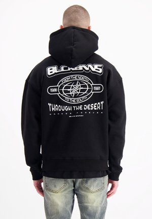 Zwarte hoodie met een grote witte grafische opdruk op de rug, featuring tekst en een kompasontwerp, gemaakt van zachte, gestructureerde stof.