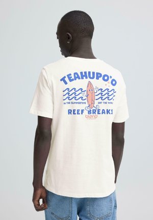 Mand iført hvid T-shirt med tegneserie surfbræt, blå bølger og teksten "TEAHUPO'O REEF BREAKS" på ryggen, set bagfra.