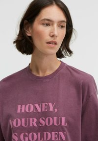 Purple cotton t-shirt featuring a round neck and bold text print in a lighter shade. The fabric appears soft with a slight texture.