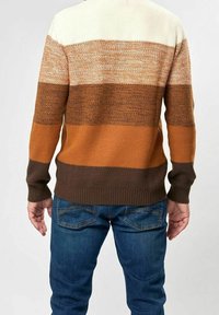 Le pull présente des rayures horizontales dans des tons crème, marron et orange, avec un tricot texturé et un ourlet côtelé, assorti à un jean en denim bleu.