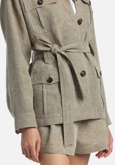 Bézs színű vászon zakó elöl gombokkal, megkötős derékövekkel, zsebfedőkkel, hozzáillő rövidnadrággal, álló személyen bemutatva.