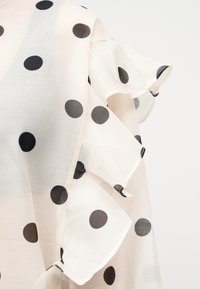 Tissu blanc à grands pois noirs avec des détails superposés et froncés le long du bord, sur un fond uni.