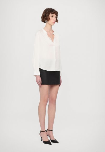 Jeune femme aux cheveux courts et bouclés portant un chemisier blanc, une mini-jupe noire et des chaussures à talons aiguilles noires, debout devant un fond blanc.