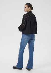 Sort top med blondeskrave og svajende ryg, parret med lyseblå flagrende jeans. Modellen har sorte ankelstøvler på. Ensfarvet hvid baggrund.