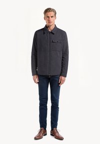 Marineblå quiltet jakke med krave og brystlomme, parret med mørke jeans og brune dressko. Glat tekstur og subtile design detaljer.