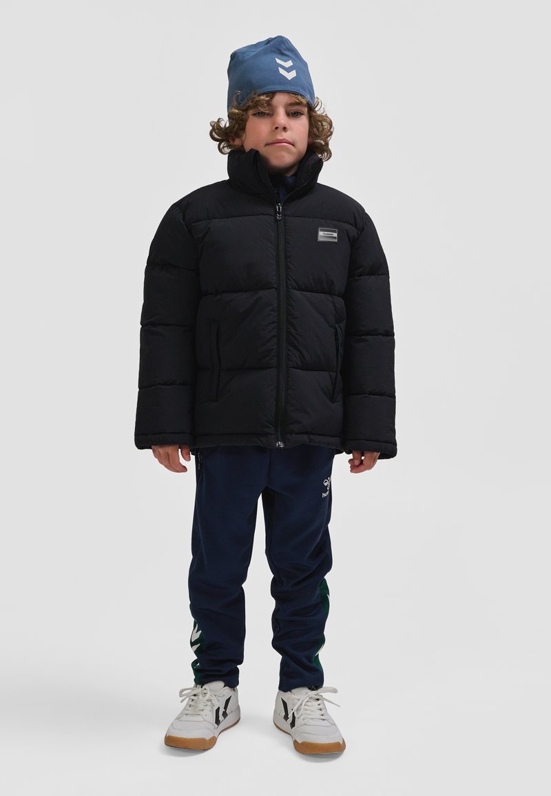 Sort dunjakke med høj krave og logo-patch, marineblå joggingbukser med grøn detalje, hvide sneakers med sorte detaljer og blå hue.