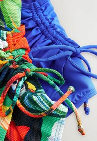 Twee paren zwembroeken: één blauwe, gemaakt van glad materiaal met een verzamelde tailleband; één kleurrijk, met een gestructureerd tropical patroon en gevlochten koord.
