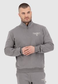 Pull en polaire gris avec un col zippé à moitié, des poignets et un ourlet côtelés, arborant un logo blanc imprimé sur la poitrine et des poches latérales.