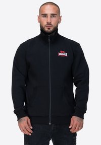 Giacca nera con zip, realizzata in tessuto morbido, caratterizzata da un colletto alto, due tasche frontali e un logo rosso ricamato sul lato sinistro del petto.