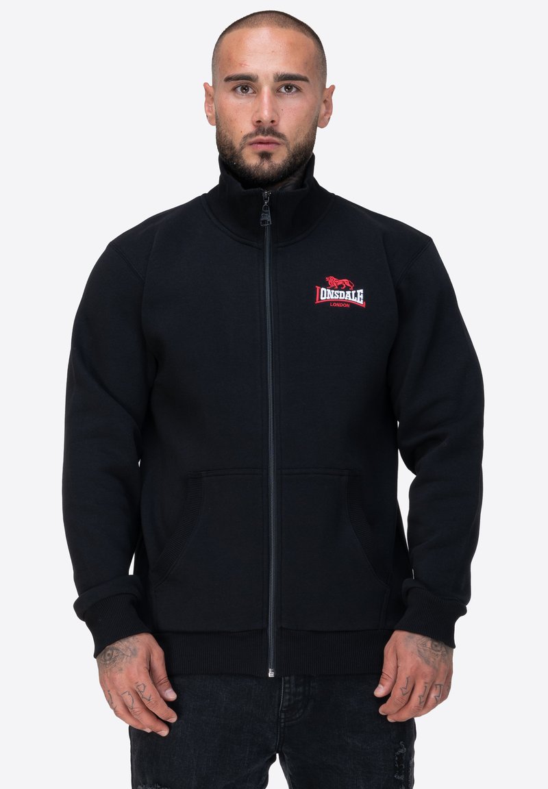 Giacca nera con zip, realizzata in tessuto morbido, caratterizzata da un colletto alto, due tasche frontali e un logo rosso ricamato sul lato sinistro del petto.