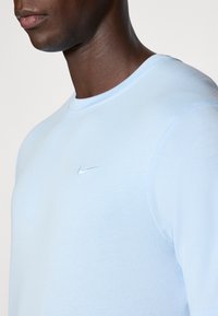 Heledad sinine pikkade varrukatega särk, mis on valmistatud pehmest kangast, ümarate kaelustega ja väikese valge tikandiga logo rinnal.