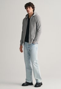 Grijze zip-up hoodie met trekkoorden, gelaagd over een zwarte shirt, gecombineerd met lichtblauwe jeans en zwarte chunky laarzen. Soepele textuur, casual pasvorm.