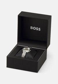 Montre argentée avec bracelet en maillons métalliques, présentée dans une boîte noire avec un intérieur noir rembourré. La boîte porte le logo "BOSS" imprimé en argent.