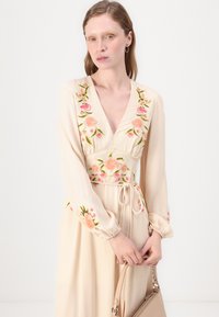 Beige mekko, jossa on kukkakirjontaa vaaleanpunaisilla ja vihreillä sävyillä. V-kaula-aukko, pitkät hihat elastisilla hihoilla ja kiristettävä vyötärö. Pitelee vaaleaa käsilaukkua.