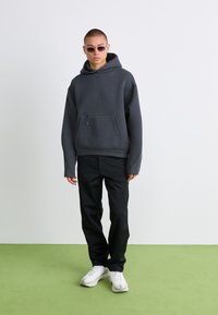 Mørkegrå hoodie med en frontlomme, lange ærmer og hætte. Sorte bukser og hvide sneakers. Model iført solbriller. Grøn gulv baggrund.