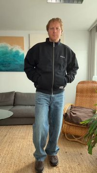 Casaco de fleece preto com fecho éclair e dois bolsos frontais, combinado com jeans azuis largos e sapatos escuros. A figura está de pé num ambiente interior, com um fundo minimalista.
