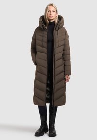 Abrigo tipo puffer largo y marr�ón con capucha, con un diseño acolchado, cierre frontal de cremallera y bolsillos laterales. Llevado sobre pantalones oscuros y botas.