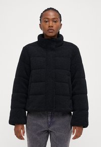 Svart, kort puffjakke med mykt, teksturert fleece-lignende materiale. Har høy krage og brede, tettsittende ermer.