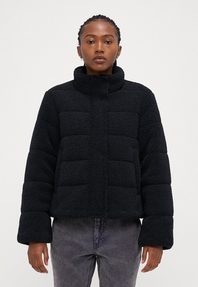 Svart, kort puffjakke med mykt, teksturert fleece-lignende materiale. Har høy krage og brede, tettsittende ermer.