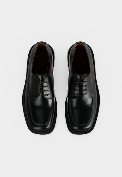 Ζευγάρι γυαλισμένα μαύρα δερμάτινα επίσημα παπούτσια με κορδόνια, τετράγωνη μύτη και ορατές ραφές σε απλό λευκό φόντο.