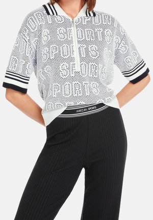 Femme portant un pull blanc zippé à manches courtes avec le texte "SPORTS" et des bordures rayées noires, associé à un pantalon noir côtelé.
