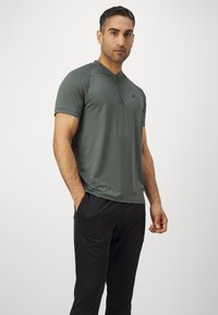 Rövid ujjú, olíva zöld ing könnyű anyagból, gallérral és cipzáras részletekkel, fekete nadrággal párosítva. Sima textúra.