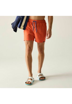 Hombre con bañador naranja con cordón azul, sandalias blancas con hebilla, sujetando una toalla doblada a rayas azul marino y beige sobre el hombro.