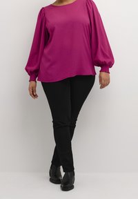 Femme portant un blouse magenta à manches longues avec des manches bouffantes, un pantalon noir ajusté, et des bottines noires, se tenant devant un fond blanc uni.