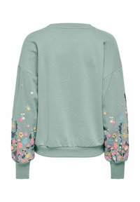 Minta zaļš krekls ar pārmērīgiem piedurknēm. Smalks ziedu izšuvums rozā, dzeltenā un zilā krāsā uz abām piedurknēm. Ribotas aproces un apakšmala.