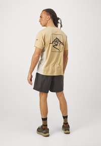 Beige og hvit kortermet skjorte med et fjellgrafikk på baksiden, kombinert med grå shorts og utendørssko.