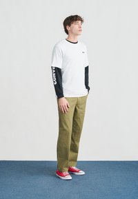 Νεαρός άνδρας στέκεται σε μπλε χαλί φορώντας λευκό και μαύρο μακρυμάνικο πουκάμισο με στρώσεις, χακί παντελόνι και κόκκινα αθλητικά παπούτσια, κοιτάζοντας στο πλάι.