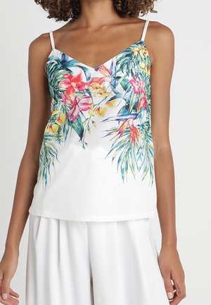 Kobieta w białej bluzce bez rękawów z kolorowym tropikalnym nadrukiem kwiatowym oraz białych spodniach z szerokimi nogawkami na jednolitym tle.