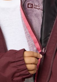 Burgunderfarbener Jacke mit Reißverschluss, ausgestattet mit einem weichen lila Fleece-Futter und rosa Akzenten. Sichtbare Hand, die den Reißverschluss greift.