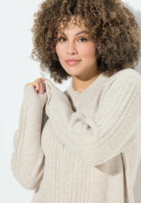 Maglione beige a maglia con texture a coste e dettagli a treccia. Maniche lunghe con polsini ripiegati, realizzato in un materiale morbido e accogliente.
