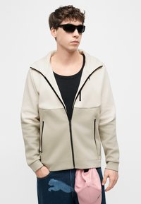 Νέος άνδρας που φοράει μπεζ φούτερ με φερμουάρ, μαύρα γυαλιά ηλίου, μαύρο πουκάμισο και μπλε τζιν κρατώντας ένα ροζ καπέλο μπέιζμπολ σε λευκό φόντο.