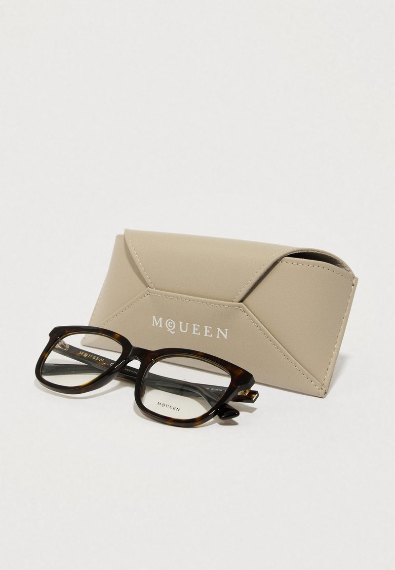 Lunettes à ecaille avec montures rectangulaires et verres transparents à côté d'un étui en similicuir beige avec le texte embossé "MQUEEN".