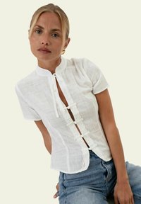 Femme aux cheveux blonds portant une blouse blanche à manches courtes avec bordure en dentelle et liens, associée à un jean bleu, debout devant un fond uni.