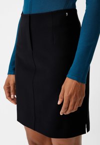 Jupe noire au design ajusté, dotée d'une fente à l'avant, d'une texture lisse et d'un logo discret sur la taille, associée à un haut bleu à manches longues.