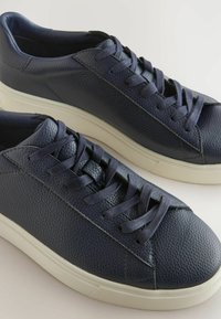 Tumedat sinise nahast sneakersid tekstureeritud viimistlusega, ümarate varvastega ja lame valge kummitald. Omavad vastavaid paelu ja minimaalset õmblustäid.