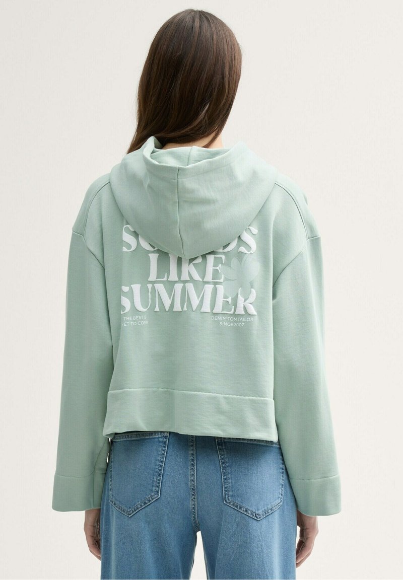 Femme aux longs cheveux bruns portant un hoodie court vert menthe avec l'inscription "Sounds Like Summer" et un jean bleu, dos à la caméra.