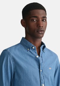 Lyseblå denimskjorte med knapp- ned krage, lange ermer og hvite knappdetaljer. Har en liten logo på brystet.