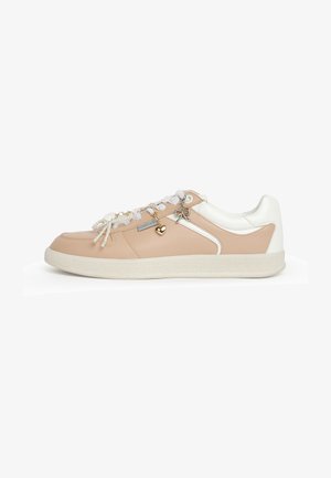 Baskets beige et blanches avec des lacets blancs, des breloques en forme de cœur perlées et dorées, et une breloque en forme de nœud métallique sur le côté.