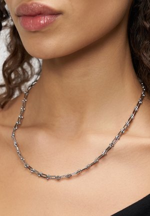 Close-up van de nek van een vrouw die een fijne zilveren ketting met kleine verbonden staafjes en kraaltjes draagt.