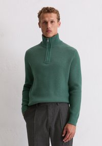 Pull-over en tricot côtelé vert avec un col haut et une fermeture éclair mi-longue. Associé à un pantalon gris sur mesure, mettant en valeur le contraste de textures et de couleurs.