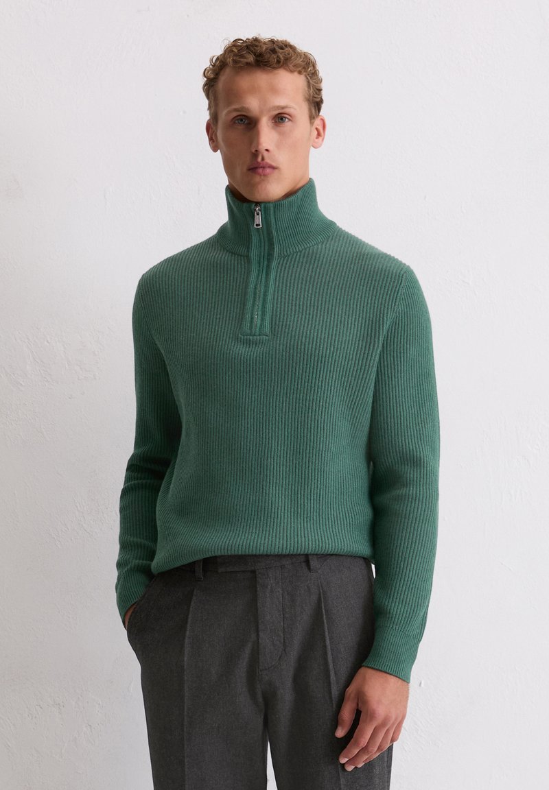 Pull-over en tricot côtelé vert avec un col haut et une fermeture éclair mi-longue. Associé à un pantalon gris sur mesure, mettant en valeur le contraste de textures et de couleurs.