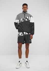 Juoda ir balta vėjinė striukė su "KANU" logotipu, tekstūruota viršutinė dalis, elastingi rankogaliai, derinama su juodais šortais ir baltais sportbačiais, demonstruojama ant modelio.