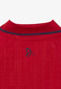 Punainen urheilu t-paita, jossa on tekstuuri-kuvio, pyöreä kaula-aukko ja tummansininen korostepanta kauluksessa. Keskellä on logo.