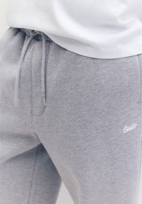Primo piano di pantaloni della tuta grigi con vita con coulisse e piccolo logo bianco sulla parte superiore della coscia, indossati con un top bianco.