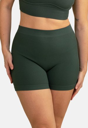 Frau trägt dunkelgrüne, gerippte Biker-Shorts mit hoher Taille und passendes kurzes Oberteil, steht mit den Händen nahe den Oberschenkeln vor weißem Hintergrund.